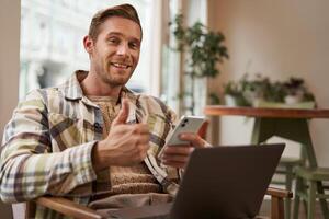 Portrait of smiling, handsome young man relaxing in coffee shop, working remotely from cafe co-working space, using laptop and smartphone, looking at camera and showing thumbs up with satisfied face photo