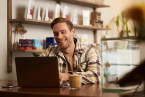 retrato de hermoso joven hombre utilizando ordenador portátil en cafetería, sentado con computadora en creativo público espacio, mecanografía en teclado con satisfecho contento sonrisa foto