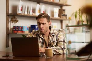 imagen de joven negocio dueño, hombre sentado en café con computadora portátil, mirando confuso a pantalla, digital nómada molesto a resolver el tarea, programador trabajando desde café tienda foto