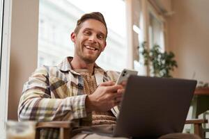 Portrait of smiling, handsome young man relaxing in coffee shop, working remotely from cafe co-working space, using laptop and smartphone, looking at camera photo