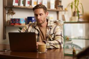 retrato de joven hombre mirando concentrado, trabajando en computadora portátil, persona de libre dedicación haciendo su proyecto en cafetería, mecanografía en teclado, Bebiendo café en trabajo colaborativo foto