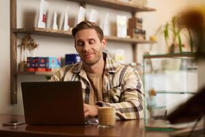 retrato de hermoso joven digital nómada, hombre trabajando en café en computadora portátil, mirando contento y satisfecho con su en línea proyecto, sentado en trabajo colaborativo café tienda foto