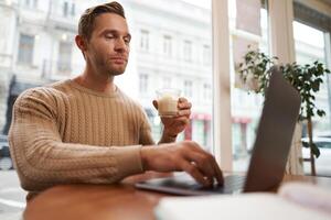 retrato de hermoso joven hombre sentado en café con ordenador portátil. digital nómada, persona de libre dedicación trabajando al aire libre desde café comercio, mecanografía alguna cosa foto