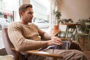 Working people and public space concept. Young handsome man, ux ui designer sitting with laptop in cafe, drinking cappuccino, looking at screen with serious face photo