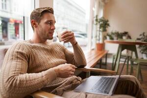 Working people and public space concept. Young handsome man, ux ui designer sitting with laptop in cafe, drinking cappuccino, looking at screen with serious face photo