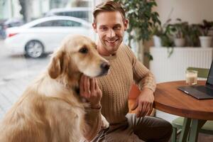 Close up portrait of smiling handsome european man with his dog in a cafe. Guy pets his golden retriever while working outdoors in coffee shop photo