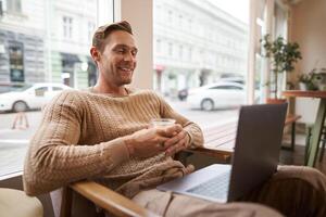 Portrait of handsome young man sits in cafe, drinks coffee and watches on laptop, looking at screen with happy smile, relaxing in co-working space photo