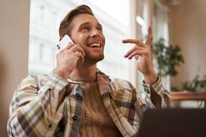 Lifestyle shot of young happy man sitting in cafe with laptop, answer phone call, talking to friend on mobile and laughing, chatting to someone with joyful face, resting in chair photo