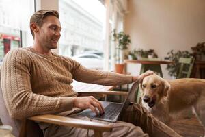 Handsome young man working in cafe with a dog, sitting on chair and using laptop, petting his golden retriever in animal-friendly co-working space photo