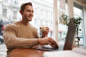 Portrait of young male businessman in cafe, drinking coffee and using his laptop, working from co-working space, sitting near the window and looking at computer screen photo