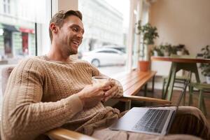 retrato de hermoso joven hombre se sienta en cafetería, bebidas café y relojes en computadora portátil, mirando a pantalla con contento sonrisa, relajante en trabajo colaborativo espacio foto