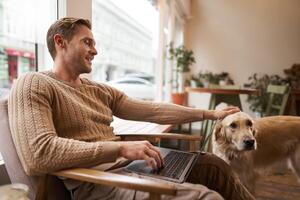 Portrait of young man, digital nomad, sitting in cafe near window, using laptop, working with his pet, petting golden retriever while sitting in chair with computer on laps photo