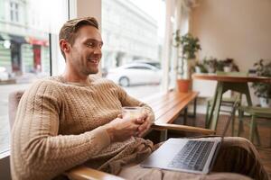 Portrait of handsome young man sits in cafe, drinks coffee and watches on laptop, looking at screen with happy smile, relaxing in co-working space photo