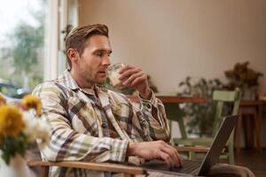 Portrait of handsome young man, working in cafe, sitting with laptop and coffee in hand, has remote job and using local co-working space instead of going to the office photo