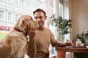 Lifestyle photo of young handsome man, entrepreneur working in a cafe on laptop while his dog sits next to him. A guy pets his golden retriever while using computer in a coffee shop