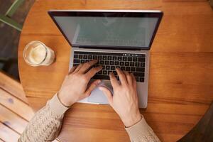 Close up portrait of male hands typing on keyboard, working on laptop in cafe, computer and cappuccino standing on wooden table in coworking space photo