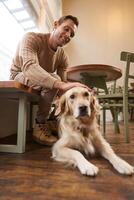 Vertical portrait of smiling handsome man with his dog, drinks coffee in pet-friendly cafe in city, touches golden retriever photo