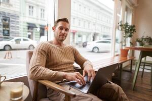 retrato de joven trabajando profesional, hombre sentado en café con computadora portátil, Bebiendo café y trabajando en línea, persona de libre dedicación utilizando trabajo colaborativo espacio a hacer su trabajo foto