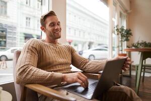 retrato de joven trabajando profesional, hombre sentado en café con computadora portátil, Bebiendo café y trabajando en línea, persona de libre dedicación utilizando trabajo colaborativo espacio a hacer su trabajo foto