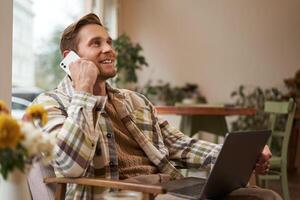 Image of young professional, cafe visitor working, sitting with laptop and talking on mobile phone to client. A guy making an enquiry buying something online, calling a friend photo