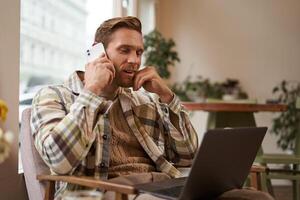 joven hombre mirando a sitio web en ordenador portátil y vocación a preguntar acerca de producto, orden alguna cosa en línea. negocio profesional sentado en café y trabajando en línea, hablando en teléfono inteligente foto