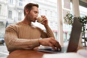 Image of handsome young businessman, man with a laptop, drinking coffee in cafe and looking at his screen, working on project in co-working space photo