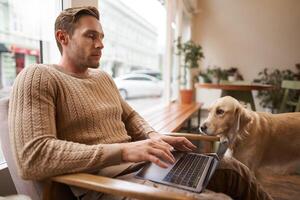 Portrait of young working man sitting in cafe, typing on keyboard while a dog looking at him. Concentrated coffee shop visitor doing his job online, sitting near window in co-working space photo