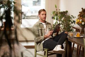 Portrait of young man sitting in cafe, spending time alone in coffee shop, reading on digital tablet, watching , listening music in wireless headphones. Lifestyle and people concept photo