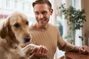 Close up portrait of beautiful golden retriever dog gives paw to a man. Smiling guy holds his pet while sits in a cafe photo