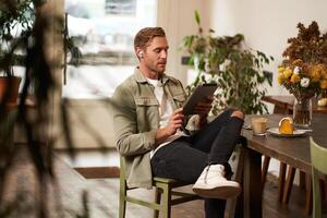 Lifestyle shot of handsome young man sitting in a cafe in front of the table, drinking coffee, wearing wireless headphones, listening to music or watching in public space, using digital tablet photo