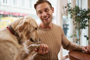 sonriente joven hombre sostiene su perro pata en mano y mira contento. concepto de mascota amigable café y Lanza libre trabajo colaborativo espacio foto