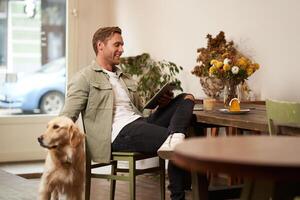 Portrait of handsome young man, cafe visitor, sitting with his dog, petting golden retriever and reading news on digital tablet, concept of lifestyle and pet-friendly public places photo