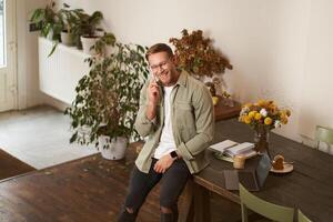 Portrait of handsome office manager, guy in co-working space, talking on mobile phone, calling someone, laughing and smiling, having pleasant conversation, sitting in office on table photo