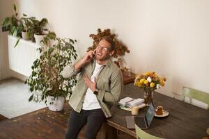 Portrait of handsome office manager, guy in co-working space, talking on mobile phone, calling someone, laughing and smiling, having pleasant conversation, sitting in office on table photo