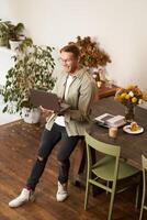 vertical retrato de hermoso contento hombre, gerente o CEO de compañía, sentado en mesa y participación computadora portátil, reír, charlando, tiene un en línea reunión con un equipo, en pie en un oficina foto