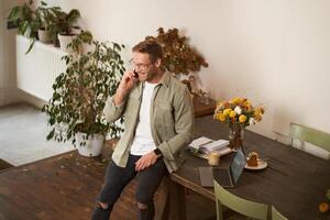 Portrait of handsome man in office, manager working in office, answering a phone call, talking to client over the telephone, sitting on table with laptop photo