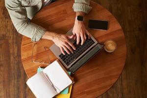 Cropped top view of male hands, man typing on laptop keyboard, working in cafe, sitting at round table in coffee shop, has phone, notebook and cup photo