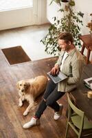 Vertical shot of handsome businessman, young man with laptop leans on table, sitting at co-working space with his dog. Young salesman checks his messages, smiling, working in an office with a pet photo