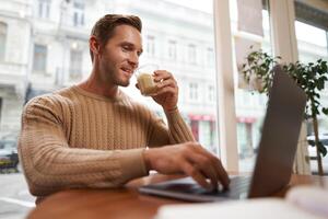 Portrait of young male businessman in cafe, drinking coffee and using his laptop, working from co-working space, sitting near the window and looking at computer screen photo