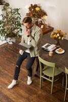 Vertical shot of handsome stylish businessman, standing near the table with laptop, thinking, working on a project on his computer, studying online, doing freelance task photo