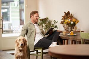 Portrait of handsome young man sits in cafe with his dog, petting golden retriever, reading news on tablet, relaxing with cup of coffee photo