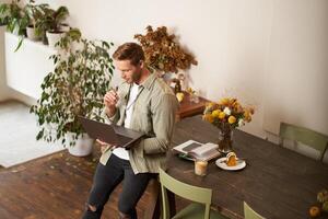 Portrait of young manager, man sits on table, holds laptop, looks concentrated at screen, reading project information, checking work data, working in office or co-working space photo