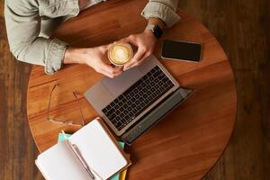 Top view. Male hands holding cup of coffee, man sitting at round table in a cafe, working on laptop, had his smartphone and notebook, drinking cappuccino photo