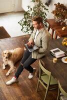 Vertical shot of handsome young man with a dog, leaning on table, holding laptop in hands, looking at screen, checking emails at work. Businessman at co-working space with his golden retriever photo