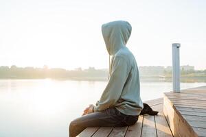 verano vacaciones por el mar, el chico se sienta meditando a amanecer, el hombre descansa en el muelle disfrutando el vista, el hipster en el capucha tiene No cabeza, el cara es cubierto foto