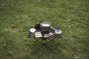 un turista mesa con comida en un caminata soportes en contra el antecedentes de césped, un plegable acampar cocina, Cocinando en el Fresco aire, compacto equipo para senderismo, comida en el mesa. foto