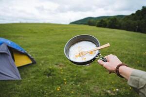 A tourist holds in his hand a frying pan with scrambled eggs, fried eggs on a hike, breakfast in nature, camping in the forest, eating scrambled eggs with a fork, a picnic in a clearing. photo