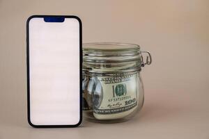 White smartphone screen mock up template in vertical position on beige background. Copy space App website advertising. Jar filled with dollars cash. Concept of Mobile application and technology business savings. photo