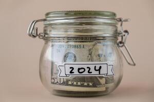 Saving Money In Glass Jar filled with Dollars banknotes. 2024 year transcription in front of jar. Managing personal finances extra income for future insecurity. Beige background photo