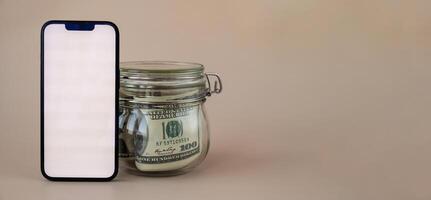 Vertical White screen on modern mobile phone in background of glass jar full of American currency dollar banknotes on beige background. Cope space for text. Advertisement for application website. Concept of money economy banks and finances photo